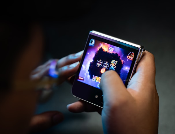 A person is holding a small electronic device, possibly a smartphone with a foldable screen, playing a video game. The game features a dark, fiery theme with icons or characters displayed on the screen.